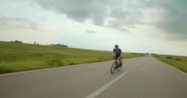 Le cycliste monte en haut. Il se tient debout sur les pédales et les tourne intensément. Entraînement à vélo. Champs en arrière-plan. 4K — Video