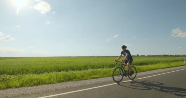 Ein Radfahrer ist auf der Straße unterwegs. Sportler trainiert auf dem Fahrrad. Grüne Wiese und im Hintergrund scheint die Sonne. 4K — Stockvideo