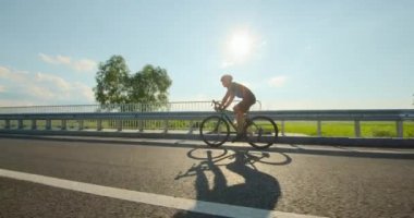 Bisikletli bir sporcu köprüden geçiyor. Bisiklet üstünde antrenman. Arka planda güneş parlıyor. 4K