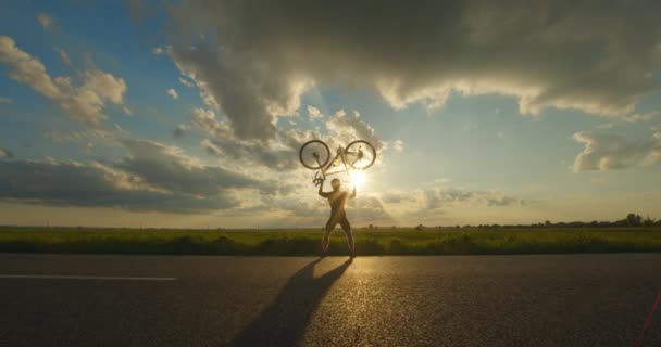 O ciclista está de pé na beira da estrada e levantando a bicicleta acima dele. Pôr-do-sol ao fundo. 4K — Vídeo de Stock