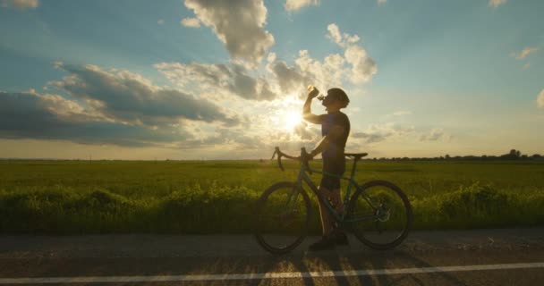 Le cycliste est debout sur le bord de la route et l'eau potable d'une bouteille Coucher de soleil en arrière-plan. La caméra zoome sur lui. 4K — Video
