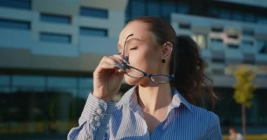 Bir iş kadını gözlüğünü çıkarıyor ve kameraya bakıyor. Yakın plan çekim. Arka planda iş merkezi var. 4K