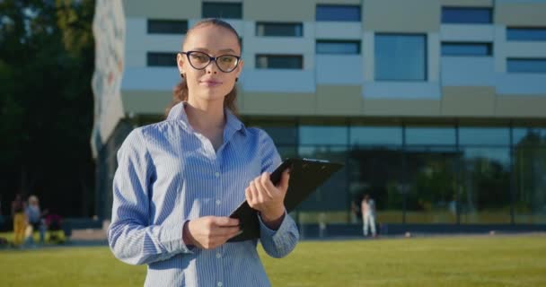 A businesswoman is standing with a folder of documents near the business center. She is smiling. Shes wearing a shirt. The sun is shining. 4K — Stock Video