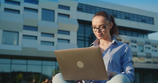 Bizneswoman siedzi na trawniku i pracuje nad laptopem. Kończy pracę i zamyka laptopa. Cieszy się blaskiem słońca. Centrum biznesowe w tle. 4K — Wideo stockowe