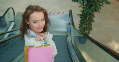 Kız yürüyen merdivenden iniyor. Kameraya bakıyor ve gülümsüyor. Omzunda alışveriş torbaları tutuyor. Alışveriş merkezinden alışveriş. 4K