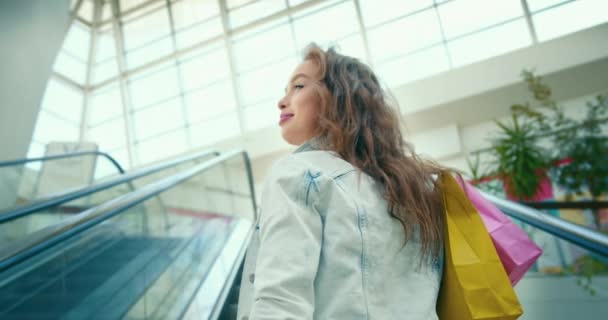 The girl is going up the escalator. She is holding shopping bags on her shoulder. She is turning and smiling. Shopping at the Mall. Shooting from behind. 4K — Stock Video