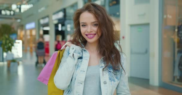 Девушка смотрит в камеру и улыбается. Она держит сумки для покупок на плече. Магазины на заднем плане. 4K — стоковое видео