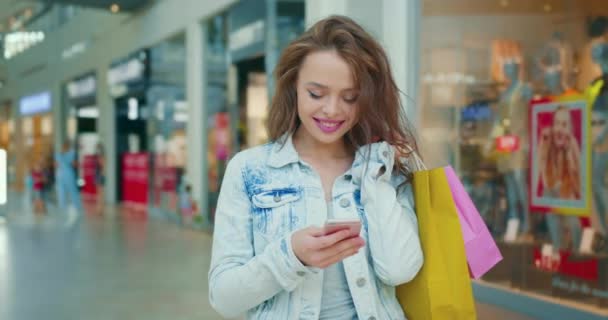 Fata zâmbește și trimite mesaje pe smartphone. Ea ține saci de cumpărături pe umăr. Magazine în fundal. 4K — Videoclip de stoc