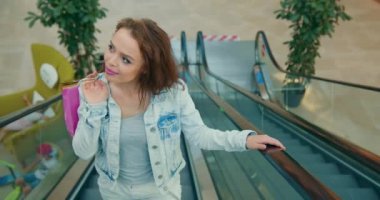 Kız gülümsüyor ve yürüyen merdivene tırmanıyor. Torbaları omzunda tutuyor. 4K