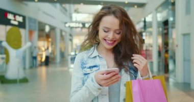 Gülümseyen bir kız telefonda mesaj atıyor ve alışveriş merkezinde yürüyor. O, elinde alışveriş torbaları tutuyor. 4K