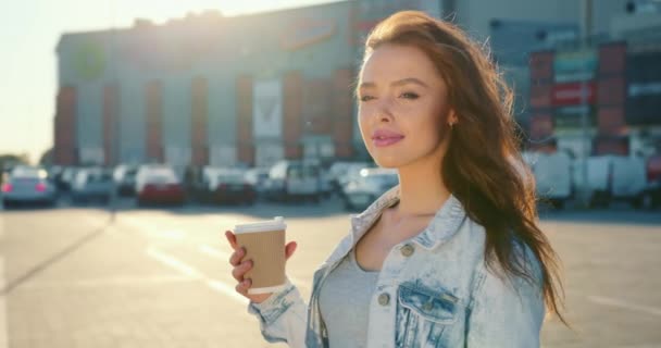 Uma menina está de pé com uma xícara de café no estacionamento de um shopping center. Ela está sorrindo. O vento sopra através do cabelo dela. Tiroteio de perto. 4K — Vídeo de Stock