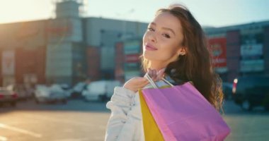 Kız dönüyor ve kameraya bakıyor. Gülümsüyor ve alışveriş torbaları tutuyor. Portre çekimi. 4K