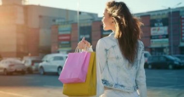 Kız alışveriş merkezinin otoparkında yürüyor ve gülümsüyor. O, elinde alışveriş torbaları tutuyor. Rüzgar saçlarını savuruyor. 4K