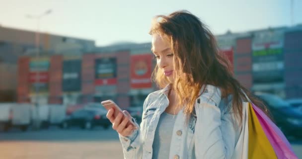 Das Mädchen schreibt SMS auf ihrem Smartphone und lächelt. Sie steht auf dem Parkplatz eines Einkaufszentrums und hält Einkaufstüten auf ihrer Schulter. 4K