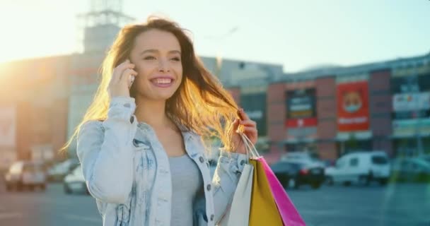 Das Mädchen telefoniert emotional. Sie lächelt und justiert ihre Haare. Sie hält Einkaufstüten in den Händen. Im Hintergrund ein Einkaufszentrum. 4K