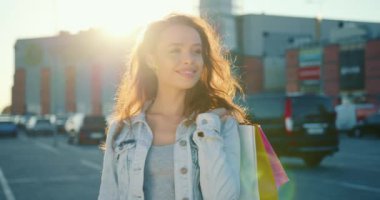 Kız ayakta ve gülümsüyor. Elinde alışveriş torbaları var. Arka planda güneş parlıyor. 4K