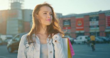 Kız bir alışveriş merkezinin otoparkında duruyor. Elinde alışveriş torbaları var. Arka planda güneş parlıyor. 4K