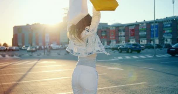 La chica está feliz y levantando las manos con los paquetes. Ella está girando y sonriendo. Está parada en el aparcamiento del centro comercial. El sol brilla en el fondo. 4K — Vídeos de Stock