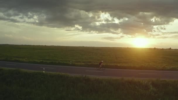 Um ciclista está andando lentamente ao longo da rodovia. A treinar numa bicicleta. Pôr-do-sol ao fundo. O drone está a contorná-lo. 4K — Vídeo de Stock