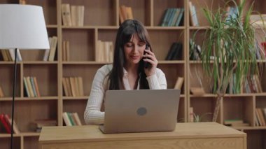 Ciddi derecede çekici beyaz bir kadın telefonla konuşuyor ve dizüstü bilgisayarına bakıyor. Stratejiyi tartışmak veya görevleri uzaktan yönetirken talimat vermek ve baskı altında sorunları çözmek.