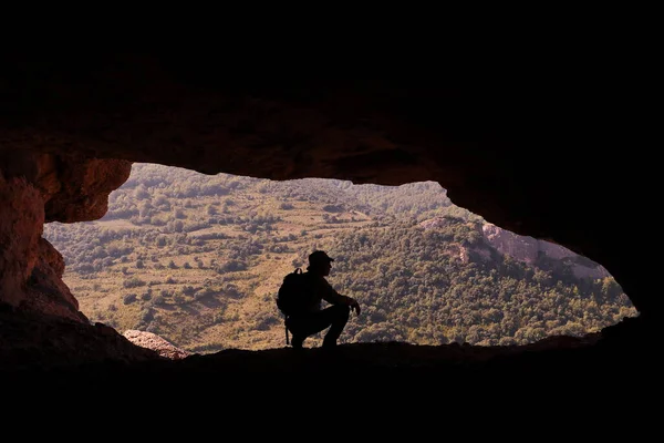 MOUNTAIN MAN WITH BACKPACK AND HAT CROUCHED AT THE ENTRANCE OF A CAVE