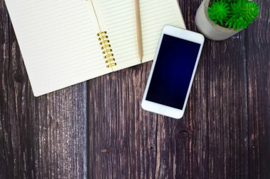 Boş, sarmal bir defter ve ahşap masa üzerinde siyah kapaklı bir cep telefonu, bitkiyle dekore edilmiş..