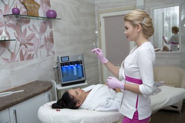 Sarı saçlı bir güzellik uzmanı hastanın yanında duruyor. Bir şırınga tutmak