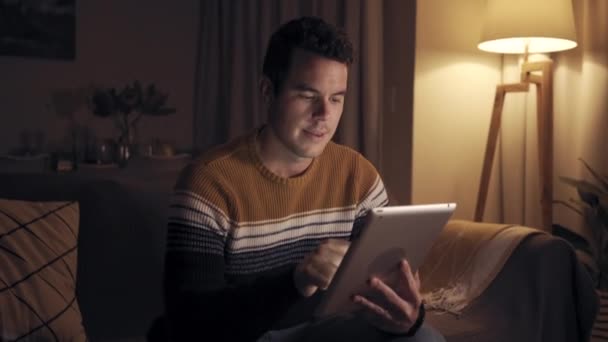 Портрет улыбающегося мужчины, сидящего на диване поздно ночью, работающего с цифровым планшетом в домашних кинотеатрах — стоковое видео