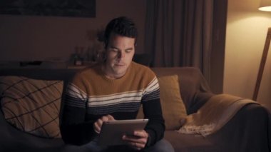 Oturma odasında gece geç saatlerde dijital tablet kullanarak kanepede rahat oturan bir adamın portresi