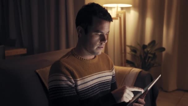 Close-up van een serieuze jonge man zittend op de Bank in zijn kamer drukke werken op digitale Tablet 's avonds laat — Stockvideo