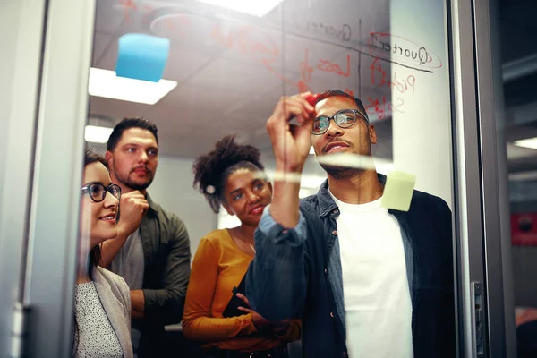 Multi equipe de negócios criativa étnica feliz olhando para o jovem escrevendo ideias em placa de vidro com marcador vermelho no escritório — Fotografia de Stock