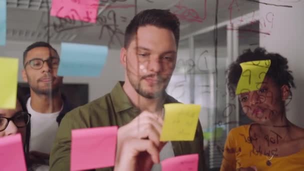 Creatieve zakenman schrijven plannen op Sticky Note op glazen wand met collega's staande in kantoor — Stockvideo