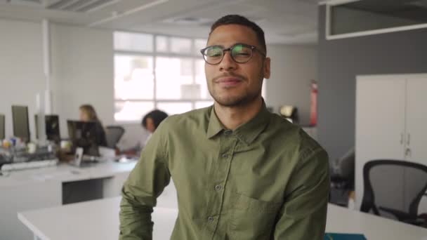 Ritratto di un giovane imprenditore afroamericano di successo che sorride e guarda la macchina fotografica seduta di fronte a un collega che lavora sullo sfondo — Video Stock