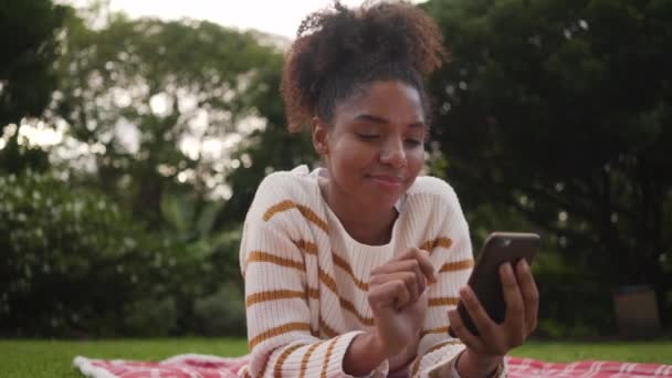 Portrait d'une jeune femme africaine couchée dans le parc profitant de messages texte sur smartphone — Video