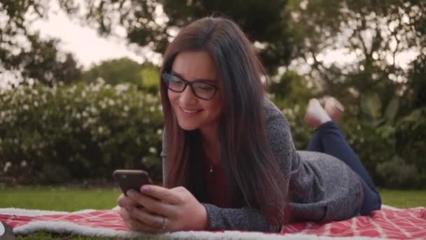 Retrato de mujer joven y sonriente relajada acostada en una manta en el parque usando un teléfono inteligente — Vídeo de stock