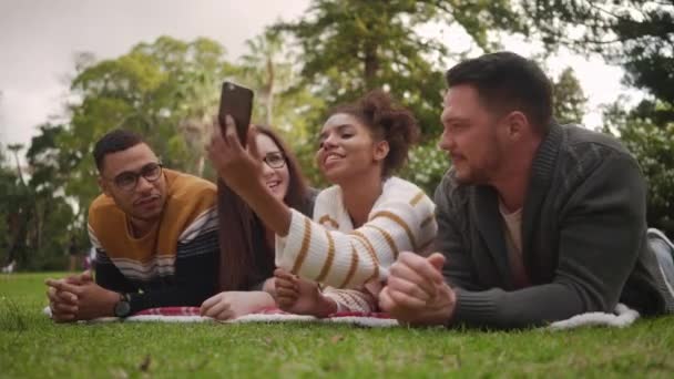 Souriante jeune femme africaine couchée avec ses amis multiraciaux dans le parc prenant selfie sur téléphone portable — Video