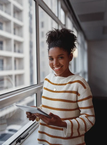 Jovem empresária brasileira sorrindo trabalhando on-line com um tablet digital enquanto estava ao lado das janelas em um escritório moderno — Fotografia de Stock