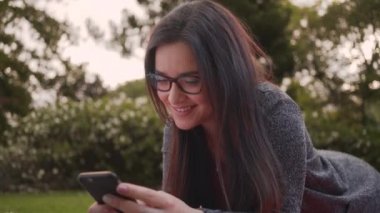 Parkta akıllı telefon kullanırken kameraya bakarak parkta yatan gözlük takan güzel bir genç kadının portresi