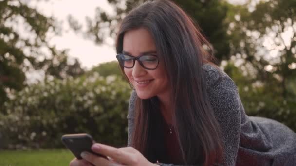 Portret van een aantrekkelijke lachende jonge vrouw liggend in Park texting berichten op smartphone — Stockvideo