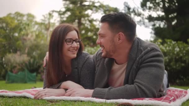 Любящая молодая пара, лежащая на одеяле над зеленой травой, целующаяся в парке — стоковое видео