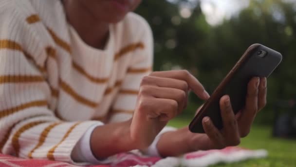 Primer plano de una joven africana acostada en el parque usando la aplicación de redes sociales en el teléfono móvil — Vídeo de stock