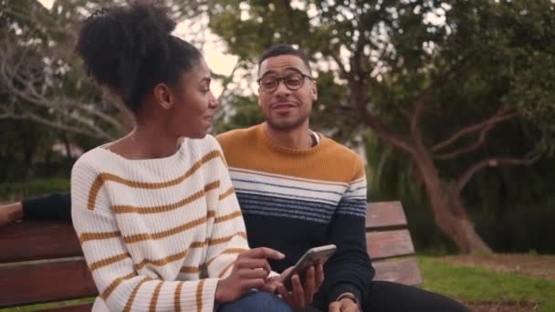 Jovem africana feliz sentada no banco com o homem usando telefone celular no parque — Vídeo de Stock