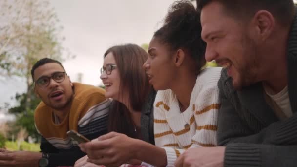 Sonriente joven africana compartiendo aplicación de redes sociales en el teléfono móvil con su amiga acostada en el parque — Vídeos de Stock