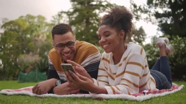 Розслаблена усміхнена африканська молода пара лежить на ковдрі над зеленою травою, приймаючи селфі на смартфон у парку — стокове відео