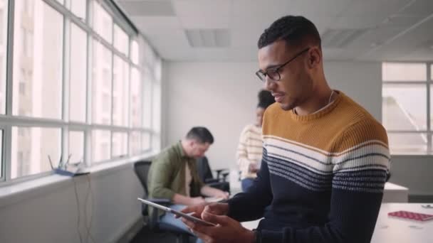 Experto joven empresario africano mirando a la cámara que sostiene la tableta digital en la oficina con colegas de equipo en segundo plano — Vídeo de stock