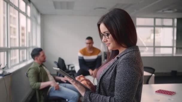 Portrait of young confident businesswoman using tablet computer smiling and looking at camera standing in front of her colleague talking in the background — Stock Video