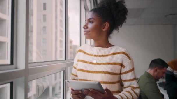 Retrato de uma jovem africana pensativa sorridente e moderna usando tablet digital em pé na frente de seu colega sentado no fundo — Vídeo de Stock