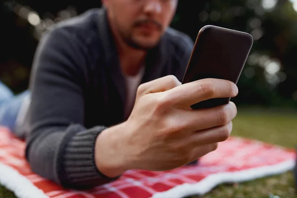 Primer plano del hombre ling en la manta sobre el césped usando el teléfono inteligente en el parque — Foto de Stock