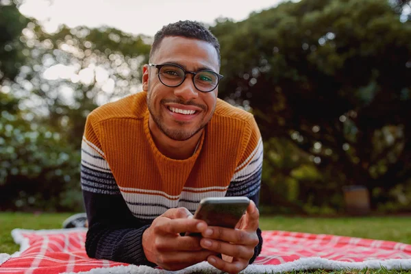 Портрет усміхненого афроамериканця, який лежить на ковдрі в парку, тримає мобільний телефон в руці, дивлячись на камеру посміхаючись — стокове фото