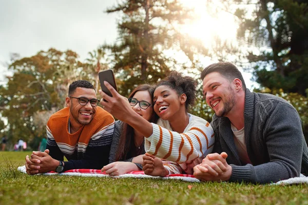 Souriant groupe multi ethnique d'amis couchés sur l'herbe verte souriant tout en prenant selfie sur smartphone dans le parc - nature très verte — Photo